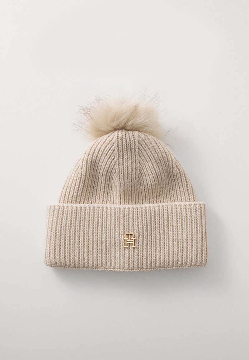 Gorro de punto beige con un diseño acanalado, que presenta un pompón de piel texturizada en la parte superior y un acento de logo dorado en el puño.