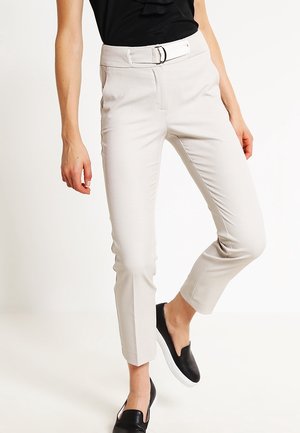 Kvinde iført tætsiddende beige bukser med hvidt bælte og sorte slip-on sko, poserer med det ene ben let bøjet.