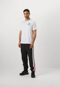 Bílé tričko s modrým logem, doplněné černými tepláky s červenými, šedými a bílými pruhy na bocích. Černé boty dokončují outfit.