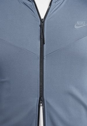Giacca zip-up in tessuto blu chiaro con una finitura testurizzata, caratterizzata da una cerniera nera e un piccolo logo nell'angolo in alto a destra.