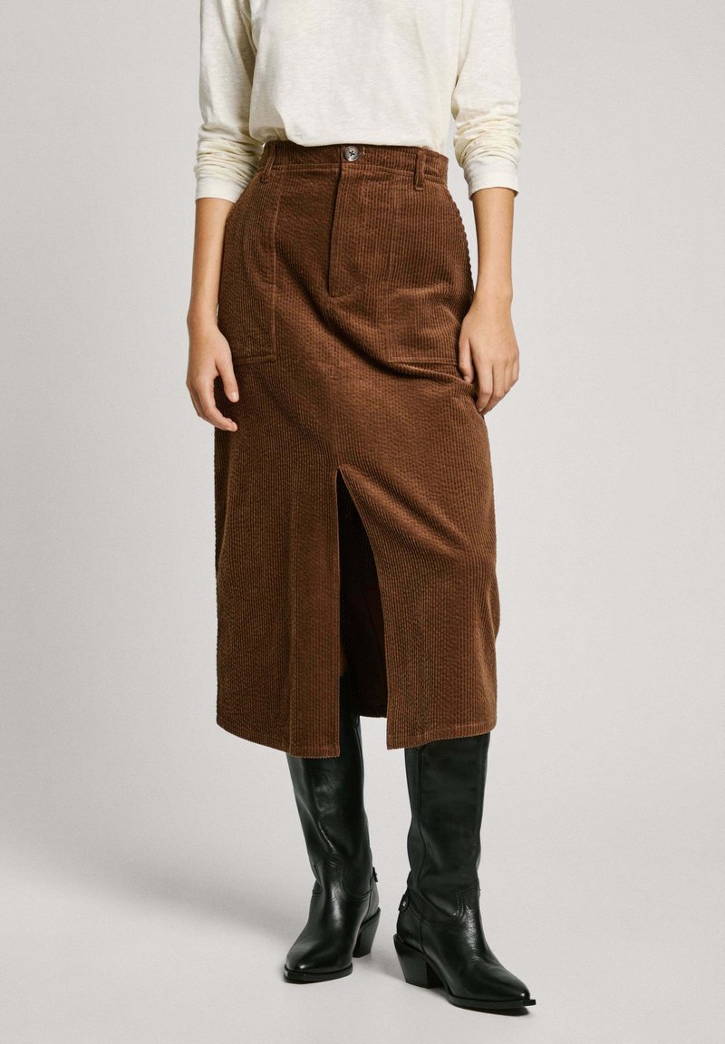 Falda midi de pana marrón con abertura frontal, con cintura alta y bolsillos, combinada con botas negras de tacón hasta la rodilla.