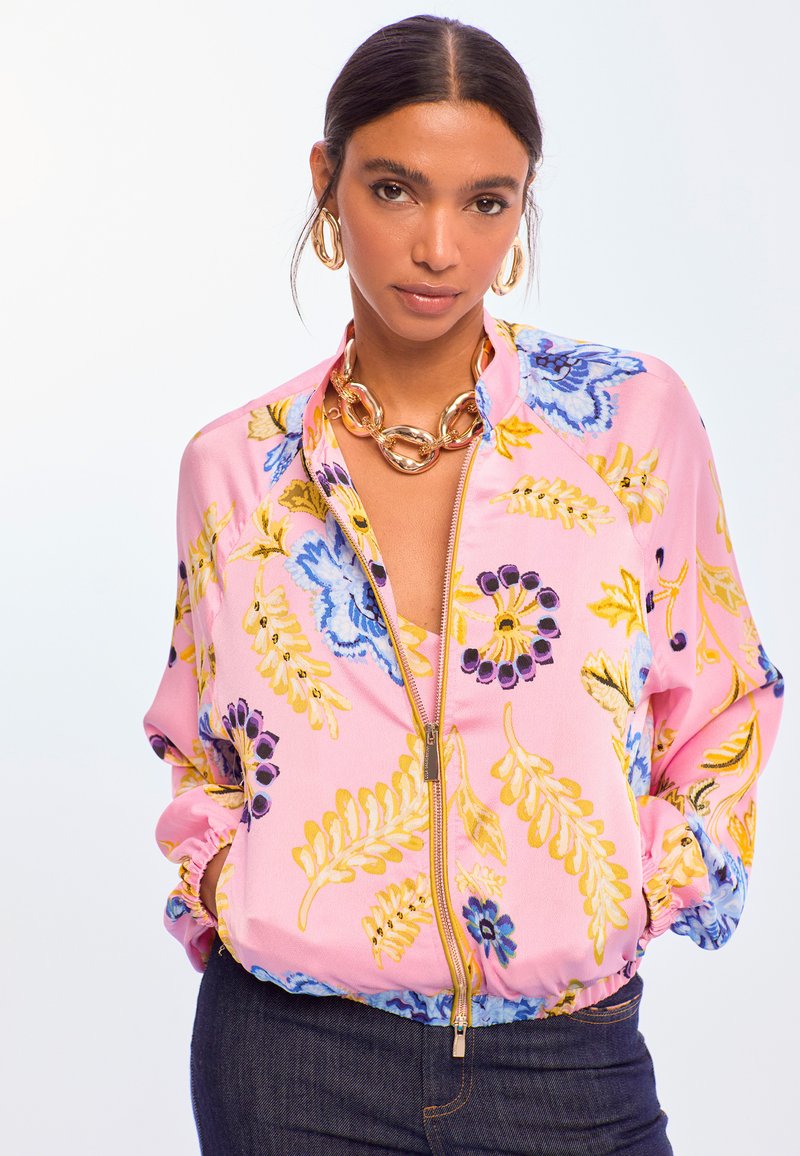 Vaaleanpunainen kukkakuvioitu bomber-takki, jossa sinisiä ja keltaisia yksityiskohtia, vetoketjusuljenta ja kootut hihansuut rentoa istuvuutta varten.