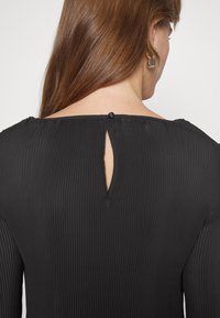 Femme aux longs cheveux bruns porte un haut noir côtelé fermé par un seul bouton dans le dos près de l'encolure.