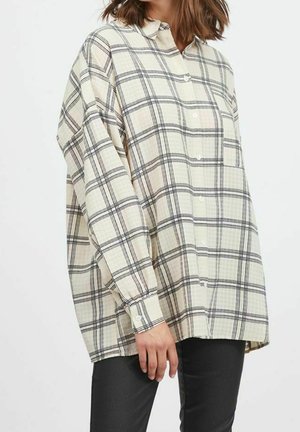 Donna che indossa una camicia oversized color crema con motivo a quadri neri, con bottoni davanti, maniche lunghe e pantaloni scuri attillati.