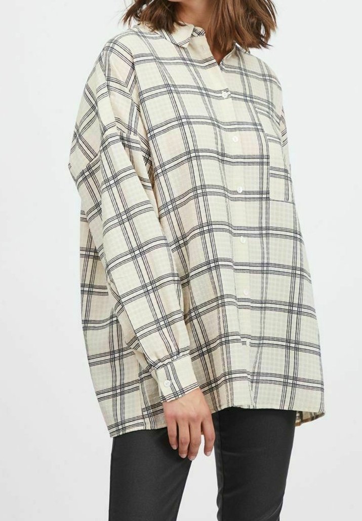 Woman wearing an oversized cream shirt with black plaid pattern, buttoned front, long sleeves, and dark fitted pants.