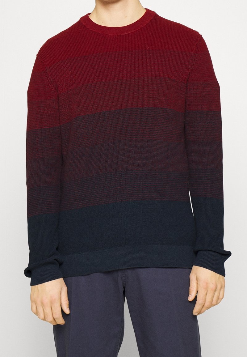 Homme portant un pull à manches longues et col rond avec des blocs de couleurs horizontaux en rouge, bordeaux et bleu foncé.