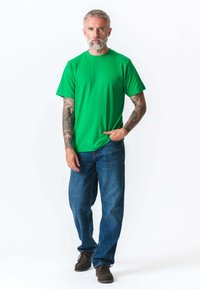 Grøn bomuld t-shirt med korte ærmer, parret med blå denim jeans. Modellen bærer også brune casual sko og har synlige tattooer på armene.