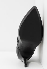 Chaussure à talon haut noire à bout pointu, positionnée avec la semelle face à l'avant sur fond blanc.