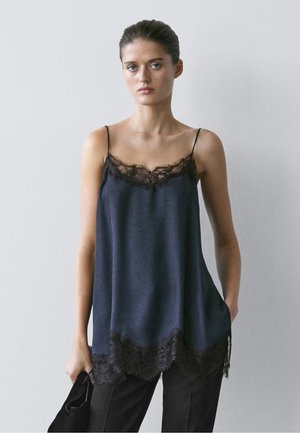 Jeune femme aux cheveux attachés, portant un caraco bleu foncé ample avec bordure en dentelle, pantalon noir, tenant un sac noir sur un fond uni.