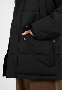 Černá péřová bunda s vycpanou strukturou. Obsahuje ploché kapsy se stiskacími knoflíky a uvolněný střih, navrstvená přes hnědý oděv.