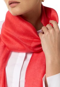 Jasnoczerwony szalik wykonany z miękkiego materiału, owinięty wokół szyi, o drobnej teksturze i subtelnym połysku, zestawiony z białą bluzką.