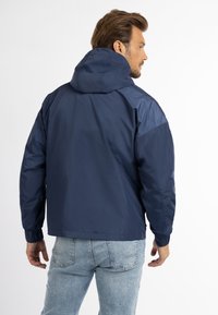 Giacca a vento navy con cappuccio, realizzata in materiale morbido e leggero. Presenta polsini elasticizzati e una vestibilità rilassata, adatta per essere indossata a strati.