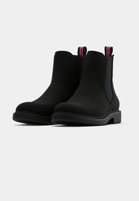 Musta Chelsea-bootit, jotka on valmistettu sileästä materiaalista. Niissä on pyöristetty kärki, matala teksturoitu pohja ja raidalliset vetoketjut punaisella, valkoisella ja sinisellä.