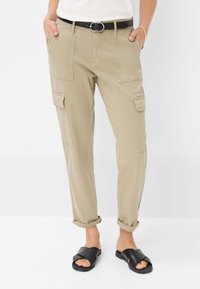 Beige cargobroeken met meerdere zakken, opgerolde manchetten en een zwarte riem. Gedragen met zwarte slipper-sandalen. Soepele stofstructuur.