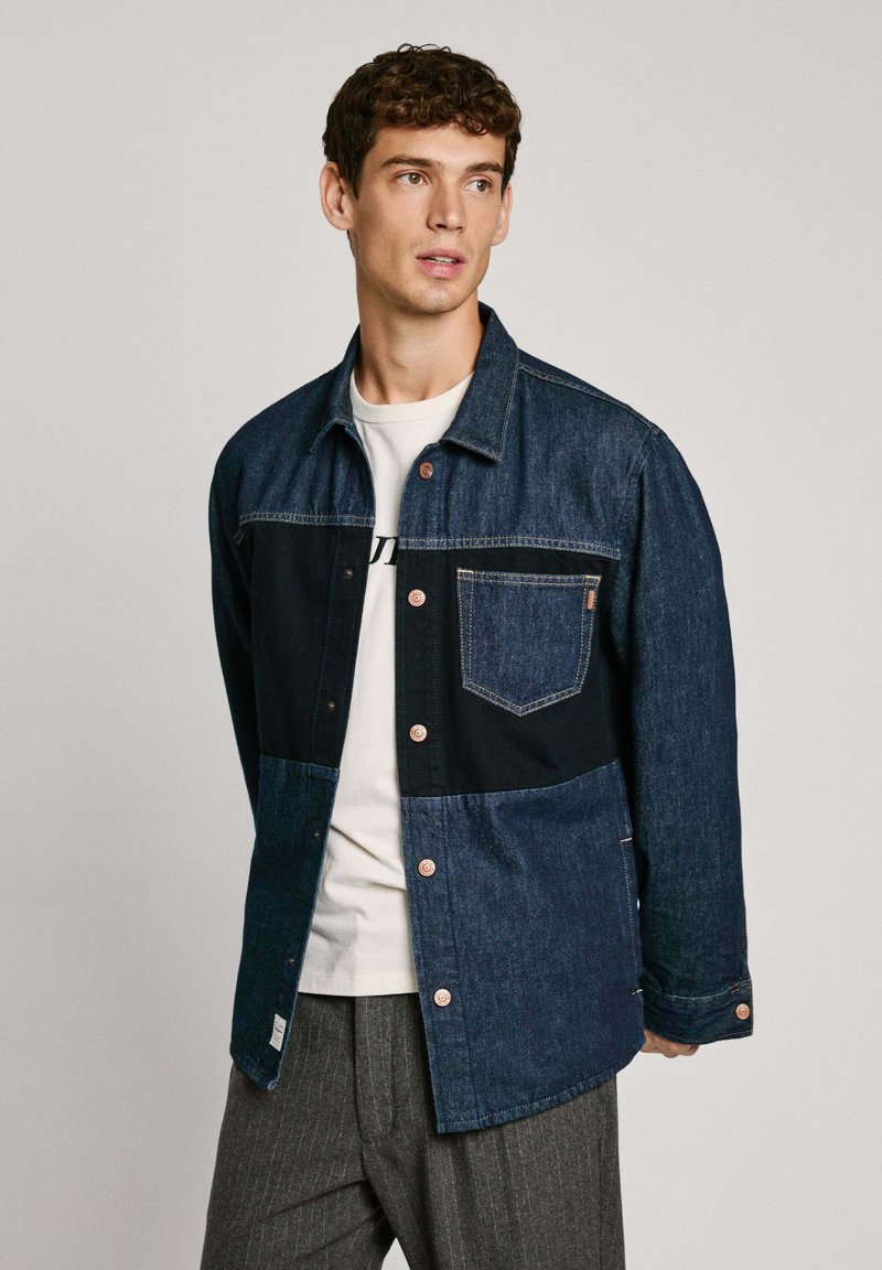 Giubbotto di denim con design bicolore: tessuto blu scuro e chiaro, caratterizzato da chiusura a bottoni, tasca sul petto e texture contrastanti.
