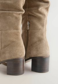 Knee-high tan suède laarzen met een licht slouchy ontwerp, een zilveren rits aan de zijkant, en een bruine houten blokhak.