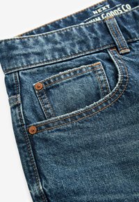 Blugi de denim într-o nuanță închisă de albastru, cu o textură uzată, având cusături contrastante, un buzunar pentru monede și un patch din piele la talie.