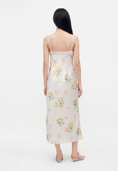 Femme aux longs cheveux noirs portant une robe nuisette blanche en satin à fleurs avec fines bretelles et talons beige, debout dos au spectateur.