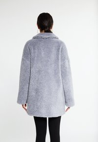 Grå fuzzy frakke med sjal-krave, oversized pasform, lange ærmer og en jævn overflade. Nederste kant er let rundet.