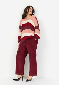 Sweter dzianinowy w poziome paski w kolorze różowym, czerwonym i kremowym; dekolt w serek. Bordowe spodnie sztruksowe; modelka w czarnych szpilkach.