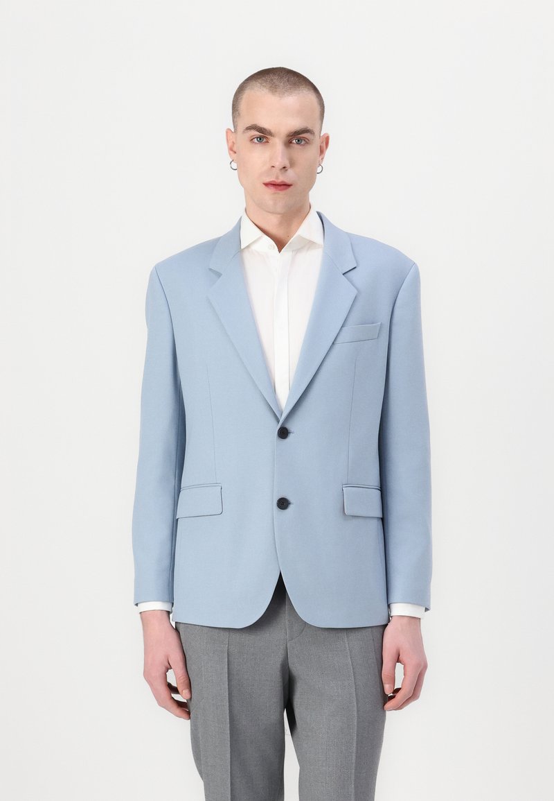 Blazer azul claro con solapas de muesca, dos bolsillos frontales y botones negros, combinado con una camisa blanca y pantalones grises. Textura de tela suave.