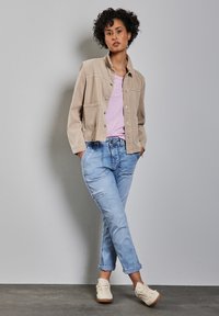 Beige bomullsjacka med knappar framtill och fickor, ljusrosa t-shirt, ljusblå uppvikta jeans och krämfärgade texturerade sneakers.