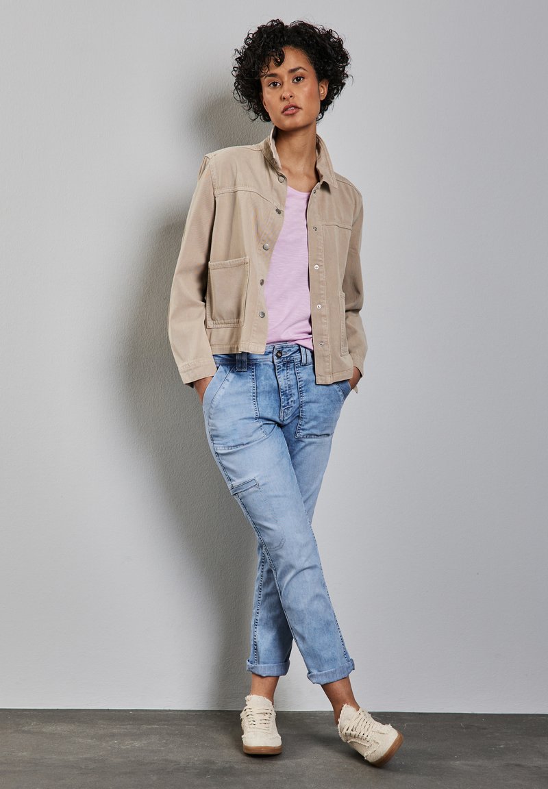 Beige bomullsjacka med knappar framtill och fickor, ljusrosa t-shirt, ljusblå uppvikta jeans och krämfärgade texturerade sneakers.