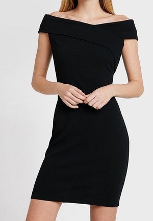 Mujer con un vestido negro ajustado, de hombros descubiertos y mangas cortas, de pie con las manos suavemente entrelazadas a la altura de la cintura.