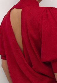 Haut en tricot rouge avec manches bouffantes et col montant. Présente une grande découpe au dos, laissant la peau visible. Le tissu a un léger effet brillant.