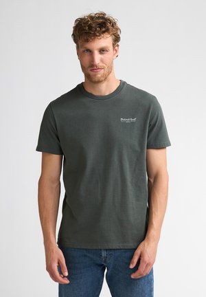 Homme aux cheveux bouclés portant un t-shirt vert foncé avec le logo Petrol-Ind et un jean bleu, debout devant un fond clair uni.