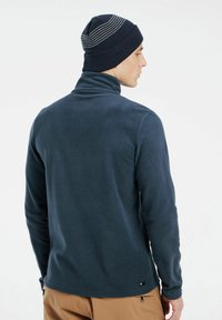 Tamsiai mėlynas fliso megztinis su aukštu apykakle, pasižymintis lygiu paviršiumi. Dėvimas su tamsiai mėlyna megztine kepure, papuošta pilkais horizontaliais dryžiais.