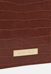 Similicuir marron avec une texture crocodile, doté d'une plaque en métal doré estampillée "ANNA FIELD". Forme rectangulaire et finition lisse.