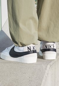 Persoon in witte en zwarte Nike sneakers met beige broek, staand op een betonnen rand buiten in het daglicht.