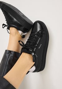 Sneaker sintetiche nere con finitura lucida, design con lacci, cerniere laterali e suola in gomma testurizzata. Presentano dettagli bianchi sul tallone.