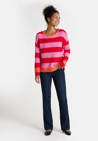 Frau steht lächelnd da, trägt einen rot-rosa gestreiften Pullover mit orangefarbenen Akzenten, dunkle Jeans und schwarze spitze Schuhe.