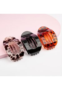 Trois pinces à cheveux rectangulaires à larges dents, motifs léopard, noir uni et écaille de tortue, disposées sur une surface rose et blanche.