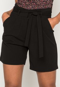 Pantalones cortos negros hechos de una tela suave con una cintura alta, pliegues frontales y un cinturón atado. Presenta bolsillos laterales para mayor funcionalidad.