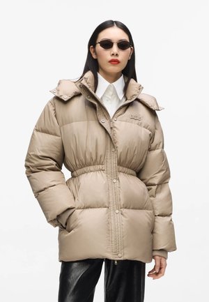 Person trägt eine beige Daunenjacke mit elastischem Bund, weißes Hemd mit Kragen, schwarze Hose und dunkle Sonnenbrille vor einfarbigem Hintergrund.