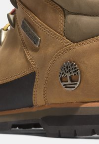 Braune Lederstiefel mit strukturiertem Finish, ausgestattet mit einem Metalllogo und Hardware-Details. Robuste Sohle mit einem kontrastierenden schwarzen Abschnitt.