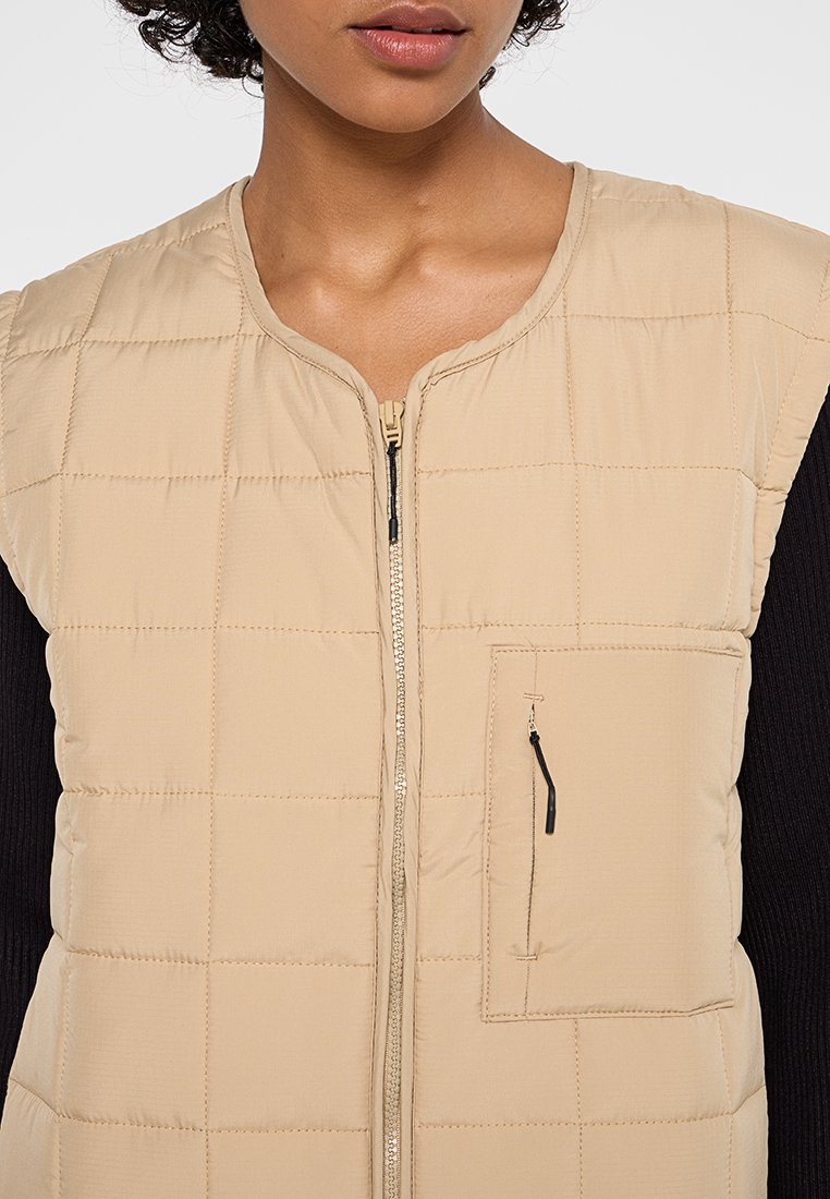 Beige Puffer-Weste mit einem gesteppten Design, Reißverschluss vorne und einer kleinen Reißverschlusstasche auf der linken Brust. Glatte Textur.
