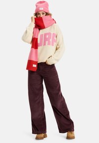 Cremefarbener Strickpullover mit pinkem "SURF"-Text, rot und rosa gestreifter Schal, dunkelviolette Cordhosen und braune Winterstiefel, ergänzt durch eine pinke Mütze.