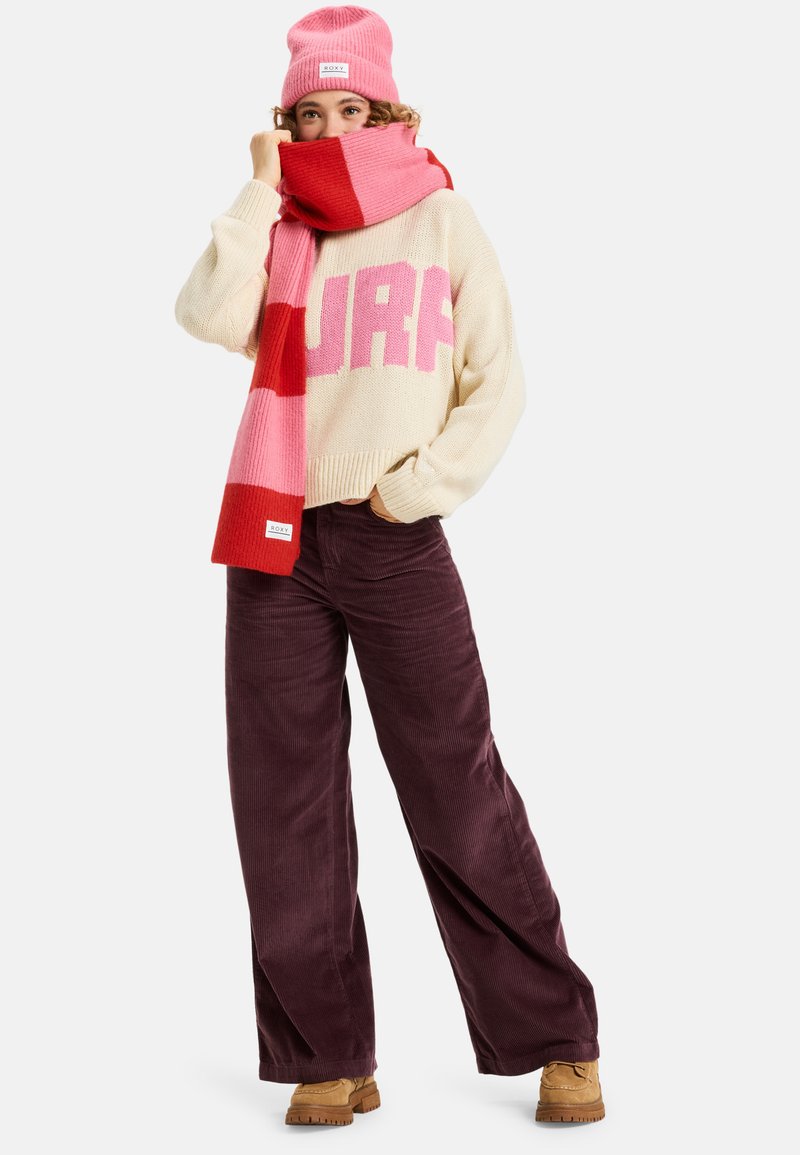 Cremefarbener Strickpullover mit pinkem "SURF"-Text, rot und rosa gestreifter Schal, dunkelviolette Cordhosen und braune Winterstiefel, ergänzt durch eine pinke Mütze.
