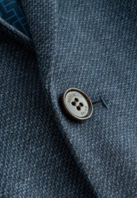 Tela texturizada de color gris oscuro con un patrón sutil, que presenta un botón redondo marrón grabado con "BAKER" en una prenda hecha a medida.