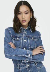 Jeune femme aux cheveux foncés mi-longs, portant une veste en jean bleue et un jean, les bras croisés, expression neutre, fond uni.