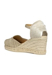 Espadrille con cuña, con parte superior de tejido beige texturizado, plantilla marrón claro y tacón de cuña envuelto en yute. Cuenta con una correa en el tobillo y una parte posterior abierta.