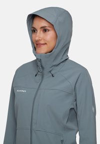 Giacca softshell di colore grigio chiaro, con zip frontale, cappuccio regolabile e branding discreto. Texture liscia con dettagli di design pratici.