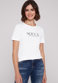 Camiseta de algodón blanca con la impresión "SOCCX TIMLESS EDITION" en negro. Manga corta y cuello redondo, combinada con jeans azules.
