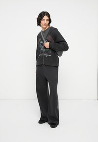 Felpa nera con zip e design grafico, abbinata a pantaloni larghi scuri. Tessuto morbido, vestibilità rilassata e dettagliata con decorazioni sottili.