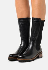Bottes en cuir noires mi-mollet avec fermeture éclair latérale et panneau texturé portées sur jambes nues, dotées d'un talon en bois empilé et d'une semelle robuste.