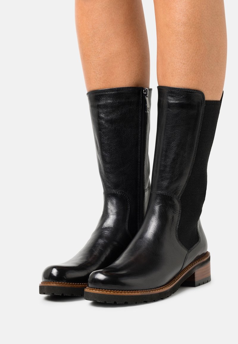 Bottes en cuir noires mi-mollet avec fermeture éclair latérale et panneau texturé portées sur jambes nues, dotées d'un talon en bois empilé et d'une semelle robuste.